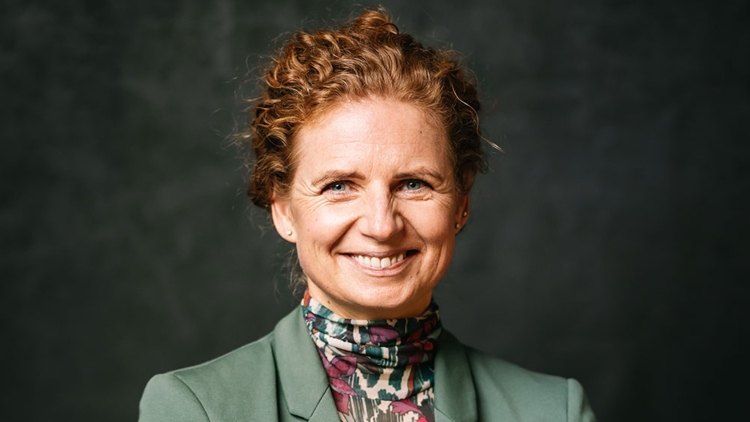 Ein Portraitfoto von Kathrin Siemer. Ein Frau, schätzungsweise 50 Jahre alt mit roten, lockigen und hochsteckten Haaren. Sie lächelt, trägt einen grünen Blazer und hat ihre Arme verschrenkt.