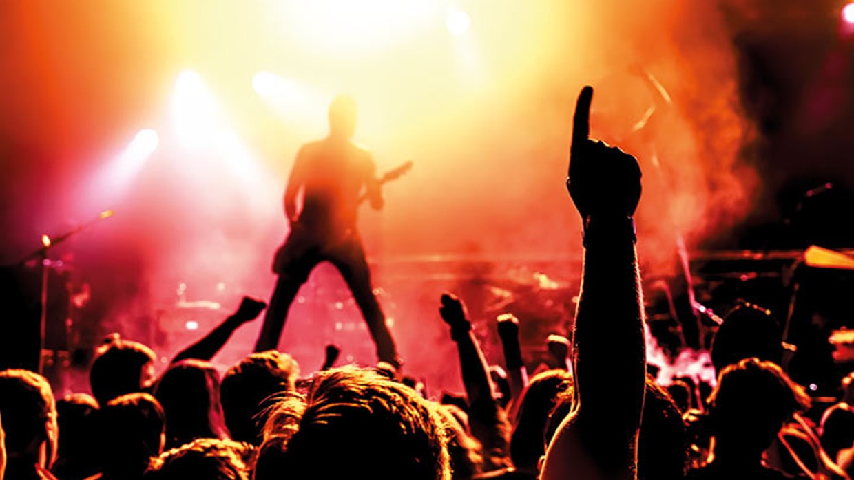 Konzertatmosphäre. Publikum im Vordergrund, im Hintergrund ist die Bühne, in gelb-orangefarbenem Licht steht ein Gitarrist mit einer E-Gitarre.